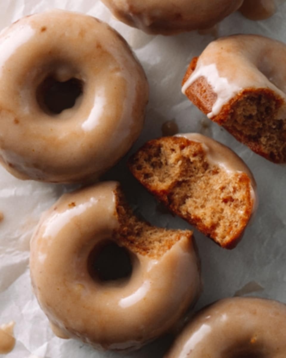 The image shows several chocolate donuts with a light brown glaze on top, arranged on a white marbled surface. One donut is broken in half, revealing a soft, moist inside that is darker brown and contrasts with the smooth, glossy glaze covering the top layer. The glaze has a slightly uneven, natural texture that looks thick and creamy. The donuts have a round shape with a hole in the middle, and their edges are slightly rough and crumbly. Photo taken with an iphone --ar 4:5 --v 7