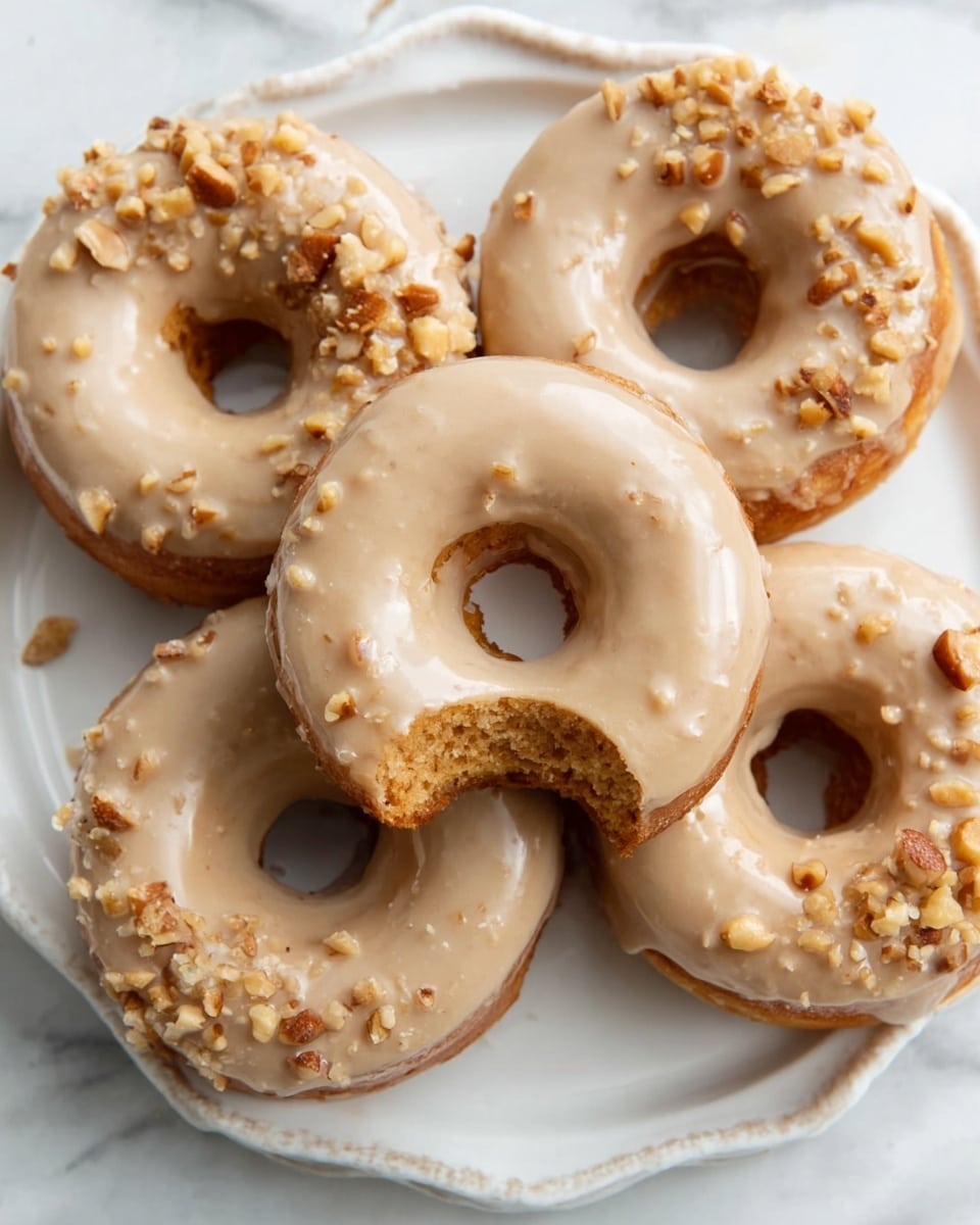 The image shows four glazed donuts on a white, round plate with a scalloped edge, resting on a white marbled surface. One donut is in the center with a bite taken out, showing a soft, light brown inside. The donuts have a smooth, light beige glaze with small bits of chopped nuts sprinkled on top, mainly clustered on the upper half of each donut. The glaze is shiny and slightly thick, covering the entire top surface. Photo taken with an iphone --ar 4:5 --v 7