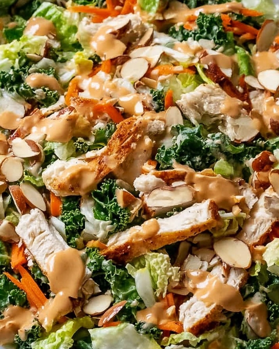 A close-up of a fresh salad showing several layers and colors, including bright green lettuce and kale leaves as the base layer. Scattered over are thin, orange carrot strips and light brown grilled chicken pieces with a slightly crispy texture. Mixed in are light green edamame beans and pale almond slices. A smooth, reddish-brown dressing is drizzled unevenly on top, adding a moist shine across the ingredients. The salad is on a white plate, placed on a white marbled surface. Photo taken with an iphone --ar 4:5 --v 7