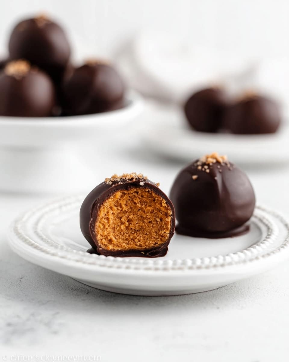The image shows three round chocolate balls on a white plate with a dotted edge, placed on a white marbled surface. One chocolate ball is cut in half, revealing a golden brown, smooth inside that looks soft and dense. The outer layer is dark and glossy chocolate with a few small chocolate sprinkles on top. In the blurred background, there are more chocolate balls on a white cake stand, also on the white marbled surface. photo taken with an iphone --ar 4:5 --v 7