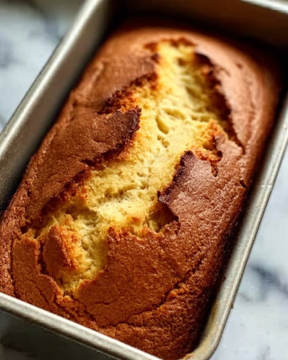 A close-up view of a freshly baked loaf cake in a silver metal baking tin, showing a cracked golden-brown crust on top with a soft, light yellow interior peeking through the crack, slightly risen with uneven texture and small air pockets, set on a white marbled surface. Photo taken with an iphone --ar 4:5 --v 7