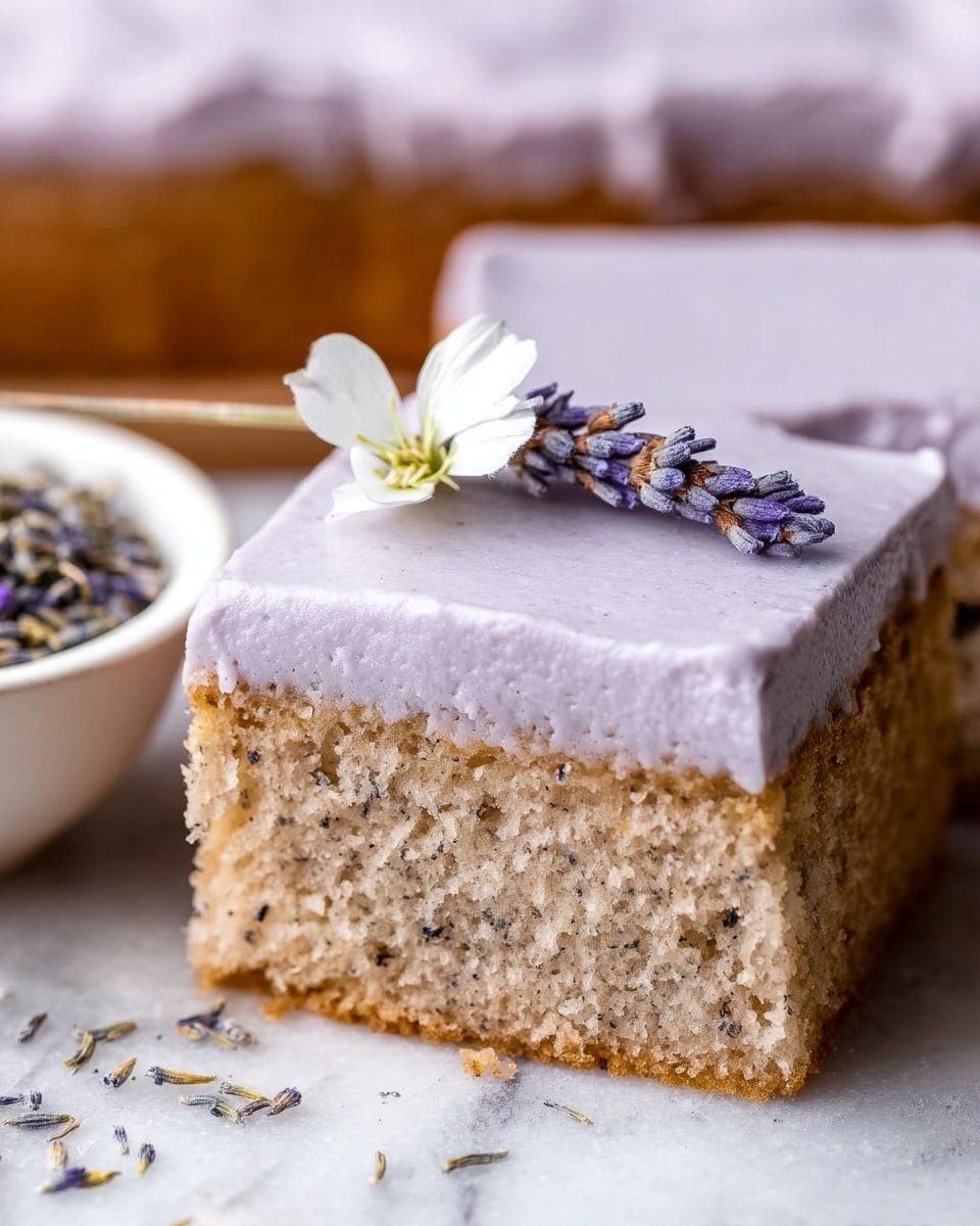 The image shows a close-up of a square slice of soft cake with two layers. The bottom layer is light brown and fluffy with small dark specks throughout, showing a moist texture. The top layer is a smooth, lavender-colored frosting that looks creamy and thick, covering the entire cake evenly. On top of the frosting rests a small, delicate white flower with light purple lavender buds nearby, adding a natural touch. The background surface is a white marbled texture. Nearby, a small white bowl holds dried lavender flowers. Photo taken with an iphone --ar 4:5 --v 7