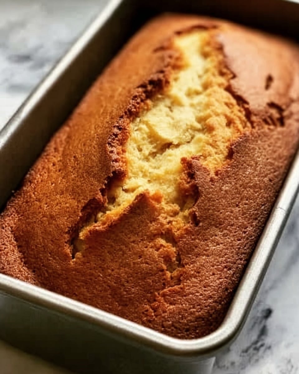 A freshly baked loaf cake with a cracked, golden brown top sits inside a metal baking pan. The cake has a rough texture with small cracks running across the surface and a deep split down the middle revealing a soft, light yellow interior. The background features a white marbled texture, highlighting the warm, inviting look of the cake. Photo taken with an iphone --ar 4:5 --v 7