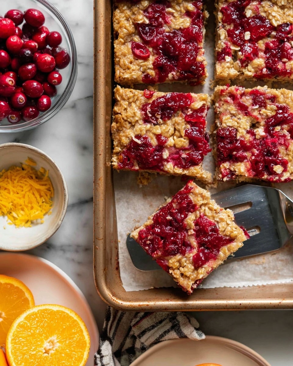The image shows three square pieces of cranberry oatmeal bars in a baking pan. Each bar has a golden-brown oat base with bright red cranberries spread throughout, giving a rough and textured look. The top layer appears slightly glossy and moist, with bits of cranberries and oatmeal visible. A spatula is partially under one of the bars, lifting it slightly. Around the pan, there is a clear bowl of whole fresh cranberries, a small white bowl with orange zest, and a white plate holding half of a sliced orange, all set on a white marbled surface. photo taken with an iphone --ar 4:5 --v 7