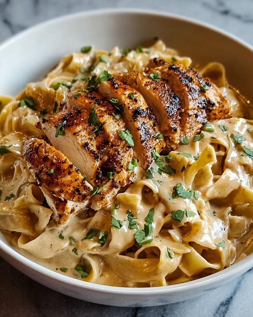 The image shows a dish with two main layers in a white bowl on a white marbled surface. The bottom layer is wide, flat noodles coated in a creamy sauce with a light tan color and a smooth, slightly glossy texture. The top layer features slices of grilled chicken breast arranged in the center, with a golden-brown crust that has a bit of char and specks of herbs. Small green herb pieces are sprinkled over the chicken and noodles, adding contrast. The overall look is warm and inviting, with good lighting that highlights the textures and colors of the food. Photo taken with an iphone --ar 4:5 --v 7