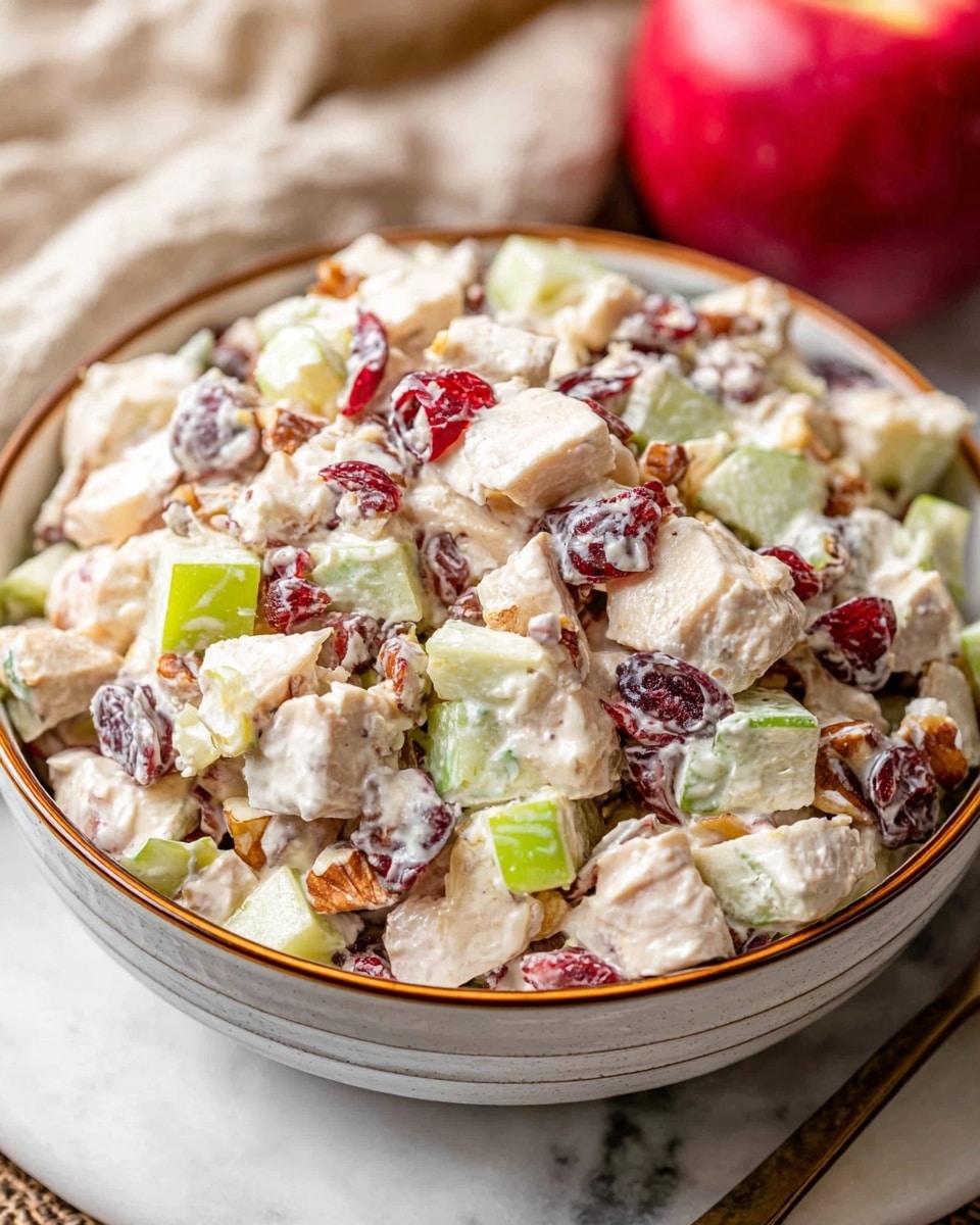 A close-up image of a creamy chicken salad in a white bowl with a brown rim, filled with small chunks of white chicken, light green celery pieces, red dried cranberries, light yellow apple cubes, and small brown nut pieces mixed in a creamy white dressing. The bowl sits on a white marbled surface, with a red apple and a light beige cloth partially visible in the background. Photo taken with an iphone --ar 4:5 --v 7