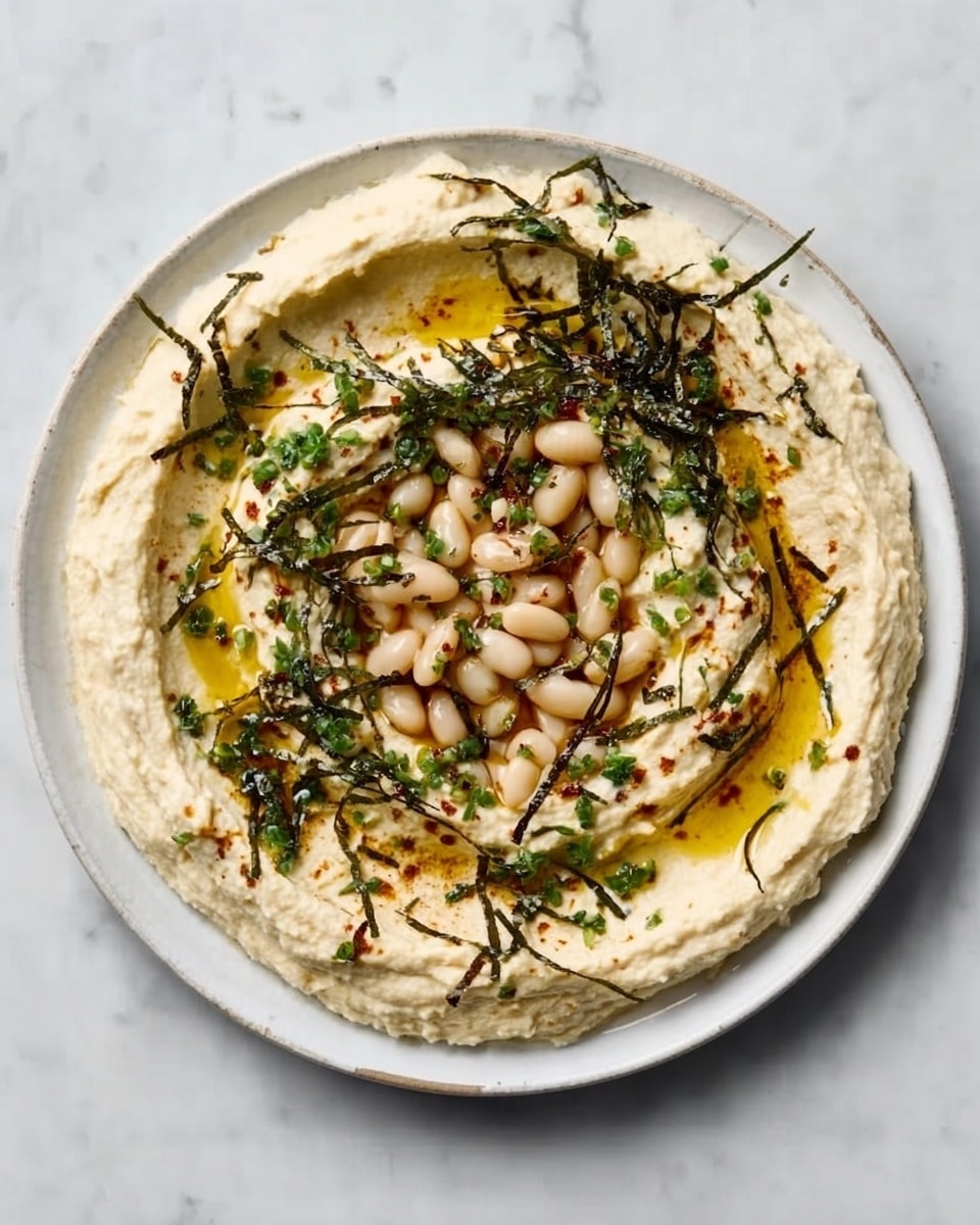 A white round plate with creamy, smooth hummus spread evenly in a circular motion covering the base, topped with small white beans scattered in the center. There are thin, dark green herbs and small golden oil pools drizzled over the hummus, adding texture and color contrast. The plate is set on a white marbled surface. photo taken with an iphone --ar 4:5 --v 7