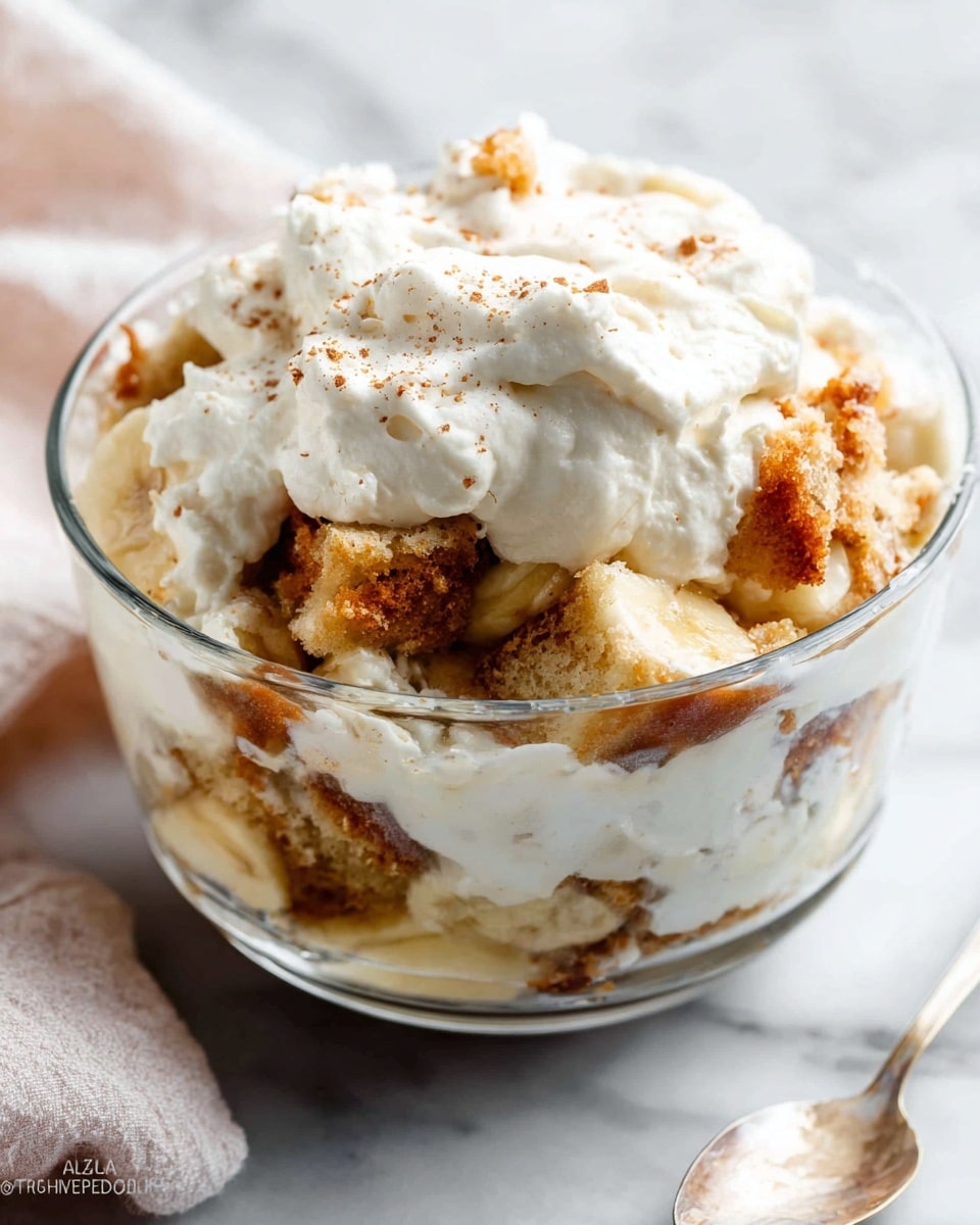 A clear glass bowl holds a layered dessert with pieces of light brown, crumbly cake mixed with chunks of soft, yellowish banana, all topped with a thick, fluffy white whipped cream that has tiny specks of brown, likely cinnamon, spread unevenly on top. The bowl sits on a white marbled surface with a corner of a light-colored cloth visible nearby. The textures range from the soft cream to the moist fruit and crumbly cake bits, creating a mix of creamy and chunky layers. Photo taken with an iphone --ar 4:5 --v 7