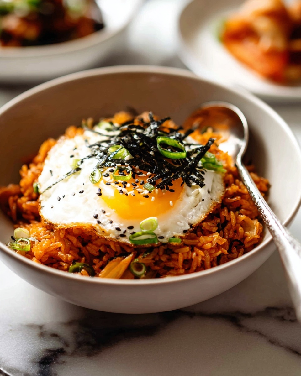 A bowl filled with a base layer of orange-red fried rice mixed with small green and beige vegetable bits, topped with a bright white fried egg with a glossy, sunny yellow yolk in the center. On top of the egg are thin black strips of seaweed scattered along with small green onion rings and black sesame seeds, adding texture and color contrast. A silver spoon rests inside the bowl on the right side. The bowl is set on a white marbled surface, with blurred dishes in the background. photo taken with an iphone --ar 4:5 --v 7