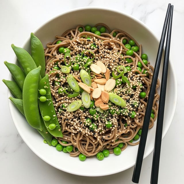 The dish is a bowl of brown soba noodles as the base layer, mixed with bright green peas scattered throughout. On top, there are thin slices of fresh green scallions and slivered pale almonds. Sprinkled all over are white and black sesame seeds, adding texture and a mix of light and dark specks. To the side of the bowl, there are vibrant green pea pods resting against the noodles. Two black chopsticks are placed on the right side of the white bowl, which sits on a white marbled surface. photo taken with an iphone --ar 4:5 --v 7