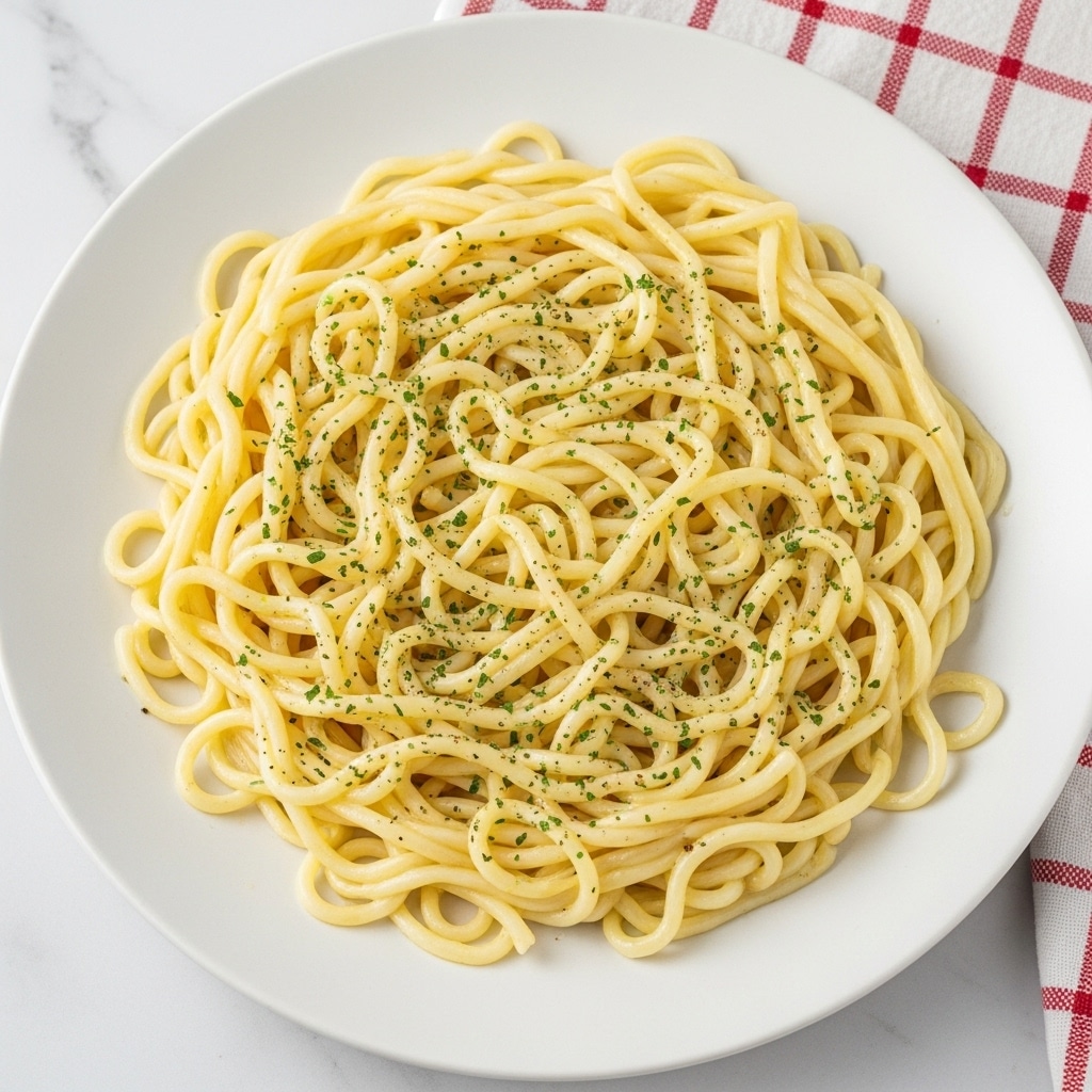 A plate filled with a single layer of glossy white noodles lightly coated in a pale yellow sauce, sprinkled evenly with small green herb flakes and specks of black pepper. The noodles have a slightly curly texture and are spread out on a plain white round plate. The plate is placed on a white marbled surface with a red and white checkered cloth visible at the top right corner of the image. photo taken with an iphone --ar 4:5 --v 7