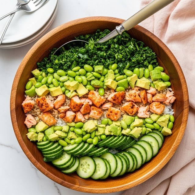 A wooden bowl filled with a colorful salad made of several layers: the base layer is thinly sliced green cucumber rounds, scattered with light green cubes of avocado, bright green edamame beans, and small pieces of cooked salmon with a slightly crispy brown edge. The texture looks fresh and varied with soft salmon flakes and crunchy cucumber slices. A large metal spoon with a light brown handle rests inside the bowl. The bowl sits on a white marbled surface next to a white plate with two forks and a pale pink cloth. photo taken with an iphone --ar 4:5 --v 7